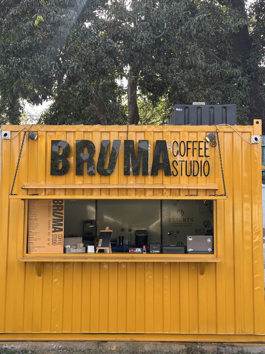 The yellow BRUMA Coffee Studio shipping-container café with its menu board and serving window, under a big tree